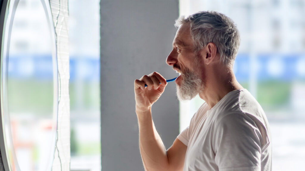 Mature man looking in the mirror and brushing his teeth to continue having good oral health after 40.