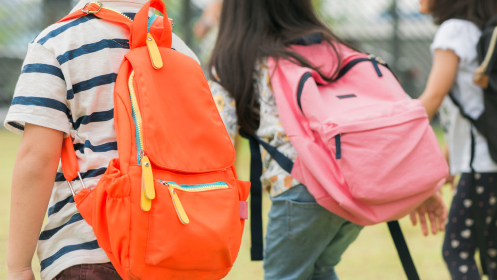 Three pupils of primary school go hand in hand. Boy and girl with school bags behind the back. Beginning of school lessons. Warm day of fall. Back to school. Little first graders. Back to school dental checkups blog article image.