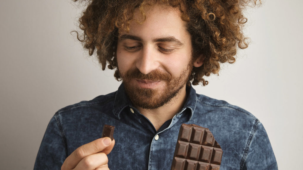 Does sugar really hurt your teeth? Image of man smiling and holding a piece of a chocolate bar ready to eat it. 