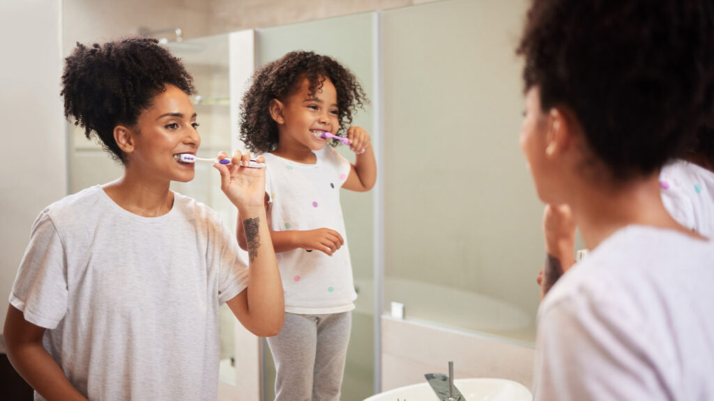 Mother and daughter brushing their teeth in the mirror after discussing when you should get a new toothbrush.